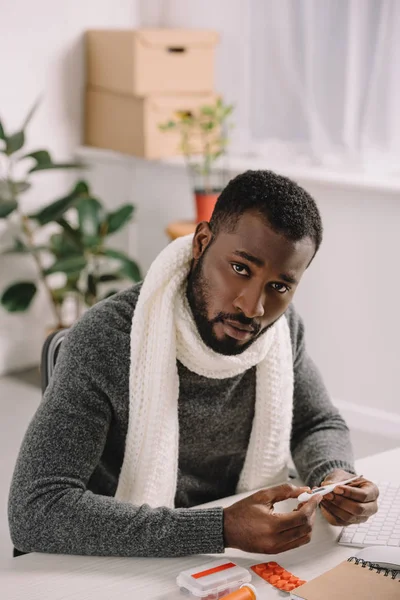 Hombre afroamericano enfermo sosteniendo termómetro electrónico en el lugar de trabajo con tratamiento - foto de stock