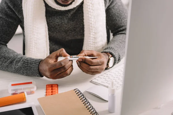 Vue recadrée du gestionnaire malade tenant un thermomètre électronique à l'espace de travail avec des médicaments — Photo de stock