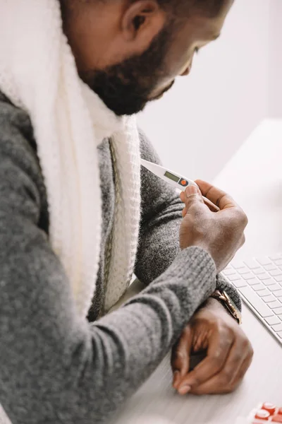 Homme afro-américain malade tenant thermomètre électronique sur le lieu de travail — Photo de stock