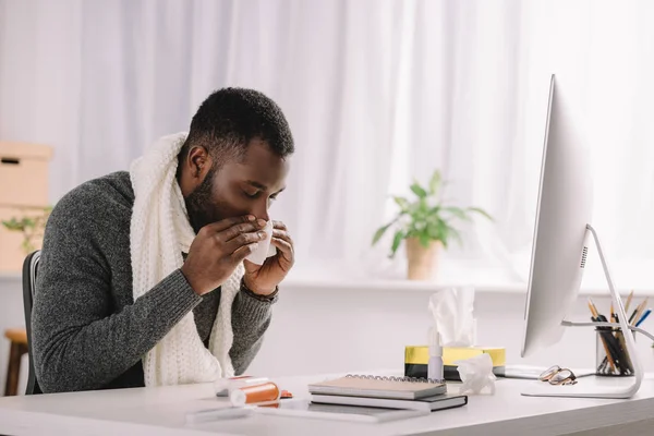 Hombre afroamericano enfermo con secreción nasal sosteniendo la servilleta mientras está sentado en el lugar de trabajo con computadora y medicamentos — Stock Photo