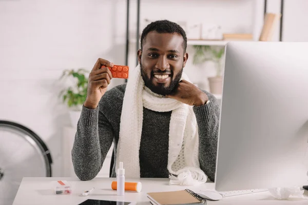 Lächelnder kranker afrikanisch-amerikanischer Mann hält Tabletten, während er am Arbeitsplatz mit Medikamenten sitzt — Stockfoto