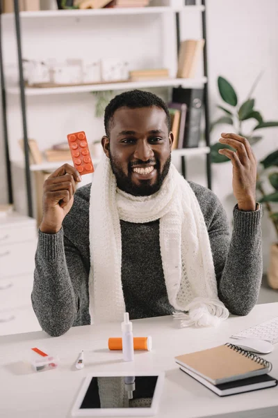 Cheerful ill african american businessman holding pills while sitting at workplace with medicines — Stock Photo
