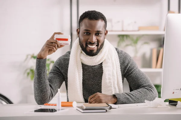Усміхнений хворий афроамериканський бізнесмен тримає коробку для таблеток, сидячи в офісі з ліками — стокове фото