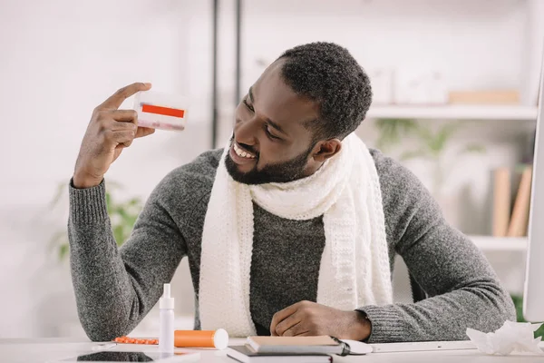 Smiling ill african american man holding pill box while sitting at workplace with medicines — Stock Photo