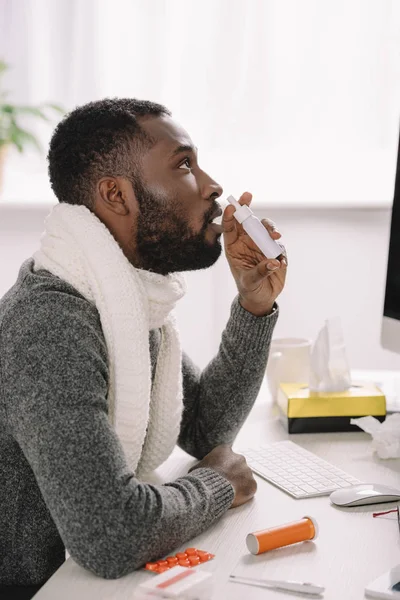 Diseased african american businessman in warm scarf with nose spray at workspace — Stock Photo