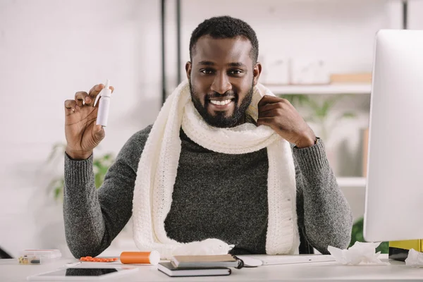Homme d'affaires afro-américain souriant froid en écharpe chaude avec spray nasal dans un bureau moderne — Photo de stock