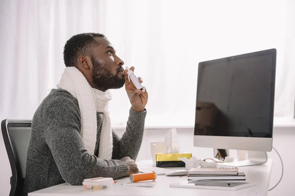 Ill african american businessman in warm scarf with nose spray at workplace with computer — Stock Photo