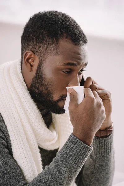 Homem americano africano frio em cachecol quente com nariz escorrendo segurando guardanapo enquanto fala no smartphone — Fotografia de Stock