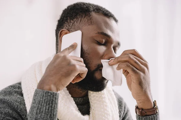 Homme afro-américain malade avec nez qui coule tenant serviette tout en parlant sur smartphone — Photo de stock