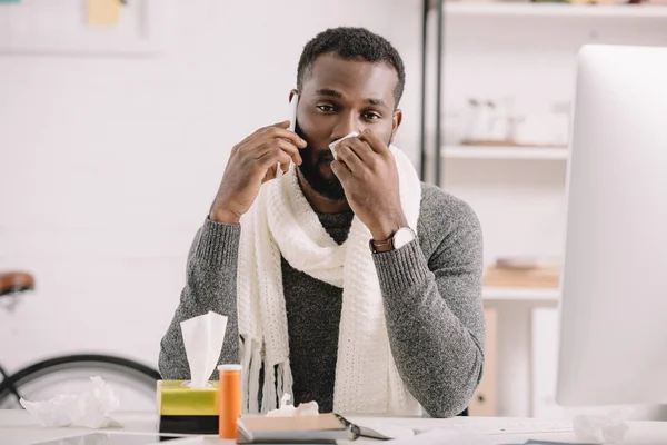 Больной афроамериканец с насморком, держащий салфетку и разговаривающий по смартфону в офисе — стоковое фото