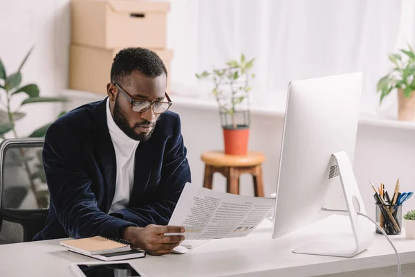 Стильний афроамериканський бізнесмен тримає документ і працює з комп'ютером в офісі — стокове фото