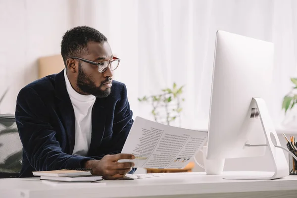 Barbudo afroamericano empresario sosteniendo documento y trabajando con la computadora en el lugar de trabajo - foto de stock