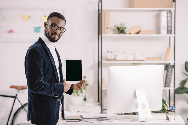 Sonriente hombre de negocios afroamericano mostrando tableta digital con pantalla en blanco en oficina moderna con computadora - foto de stock