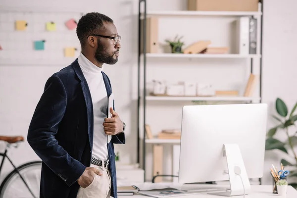 Guapo hombre de negocios afroamericano sosteniendo tableta digital en la oficina con computadora — Stock Photo