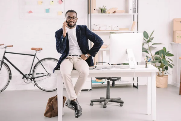 Hombre de negocios afroamericano hablando teléfono inteligente mientras está sentado en la mesa con el ordenador en la oficina moderna - foto de stock