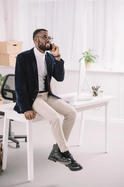 Hombre de negocios afroamericano profesional hablando teléfono inteligente mientras está sentado en la mesa en la oficina moderna con computadora - foto de stock