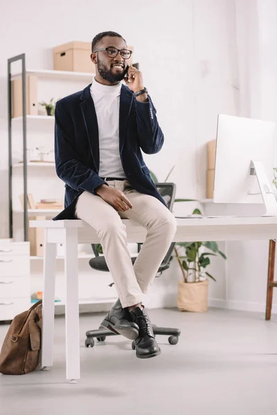 Schöner erfolgreicher afrikanisch-amerikanischer Geschäftsmann spricht Smartphone an, während er auf dem Tisch im modernen Büro sitzt — Stockfoto