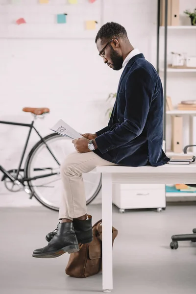 African american businessman reading newspaper in modern office with bicycle — Stock Photo