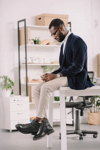 Bonito Africano americano empresário falando smartphone enquanto sentado na mesa no escritório — Fotografia de Stock