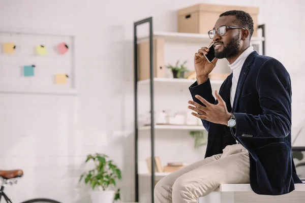 Barbuto afroamericano uomo d'affari parlando smartphone mentre seduto sul tavolo in ufficio moderno — Foto stock