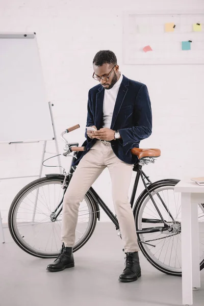 Stylish african american businessman using smartphone while leaning on bicycle in office — Stock Photo