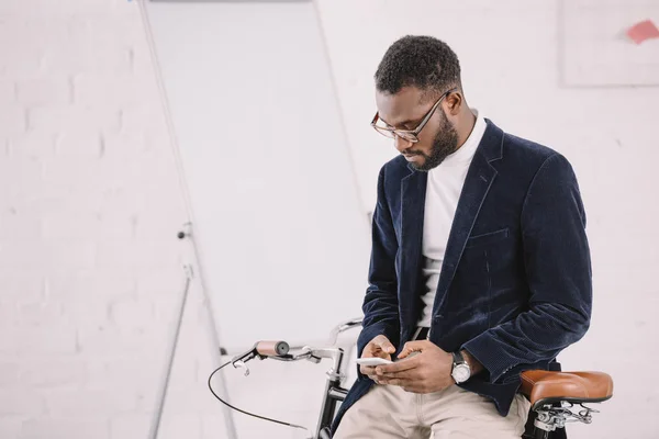 Hombre de negocios afroamericano usando smartphone mientras se apoya en bicicleta en la oficina con pizarra blanca - foto de stock