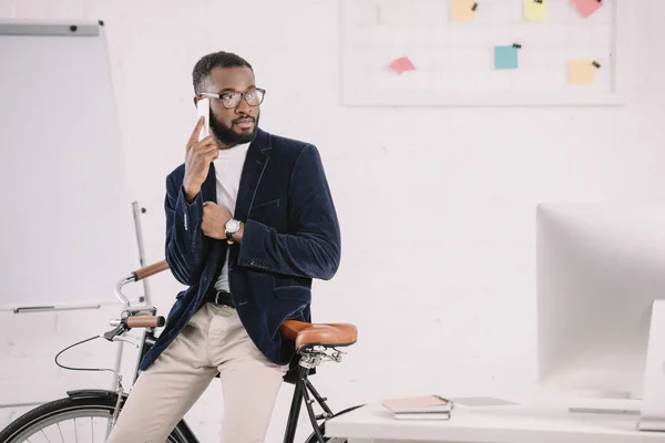 Elegante manager afroamericano che parla su smartphone mentre si appoggia sulla bicicletta in ufficio con il computer — Foto stock