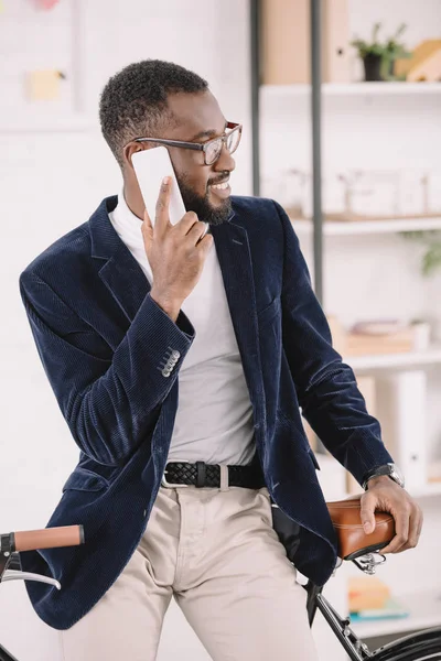 Gai gestionnaire afro-américain parlant sur smartphone tout en s'appuyant sur le vélo au bureau — Photo de stock