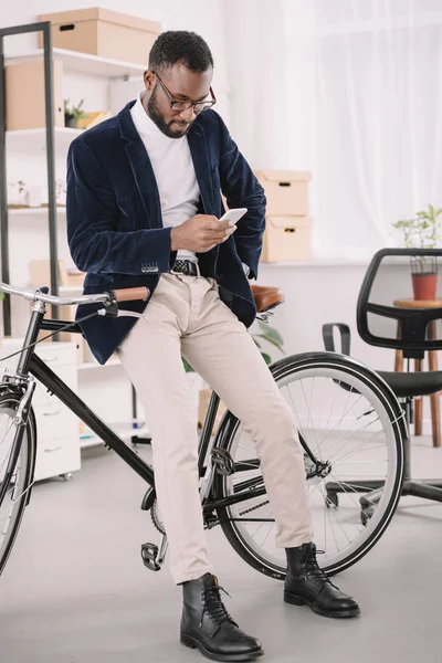 Homem de negócios americano africano barbudo usando smartphone enquanto se inclina na bicicleta no escritório — Fotografia de Stock