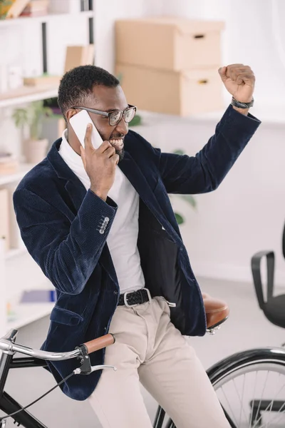 Successful african american businessman triumphing and talking on smartphone while leaning on bicycle in office — Stock Photo