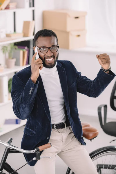 Bem sucedido empresário afro-americano falando no smartphone enquanto se inclina na bicicleta no escritório — Fotografia de Stock