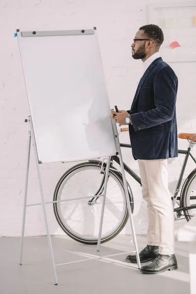 Hombre de negocios afroamericano trabajando con pizarra blanca en la oficina con bicicleta - foto de stock