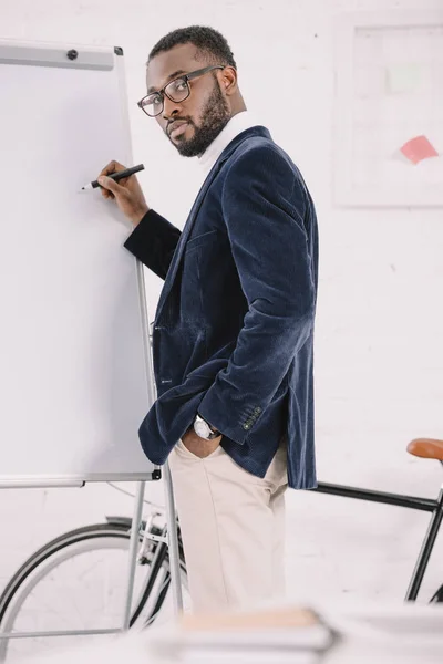 Bearded african american businessman in velvet jacket writing on whiteboard in office with bicycle — Stock Photo