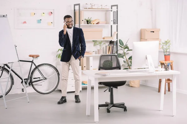 Empresario afroamericano hablando en teléfono inteligente cerca del lugar de trabajo con la computadora - foto de stock