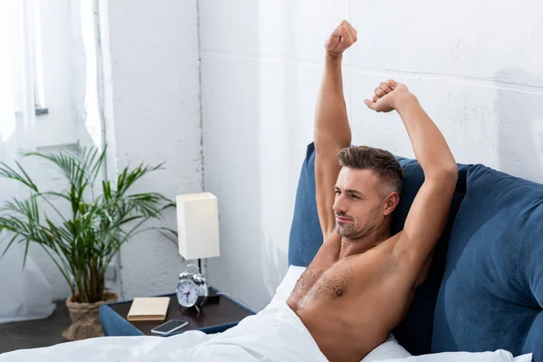 Foco seletivo do homem sem camisa com braços levantados fazendo estiramento na cama em casa — Fotografia de Stock