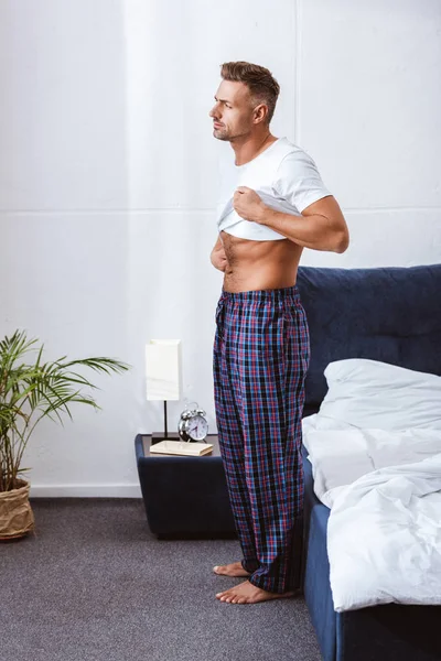 Erwachsener Mann zieht weißes T-Shirt in Bettnähe an — Stockfoto