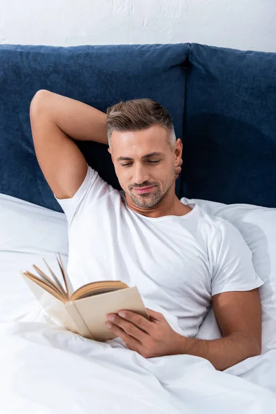 Vista de ángulo alto del libro de lectura del hombre mientras está acostado en la cama durante el tiempo de la mañana en casa - foto de stock