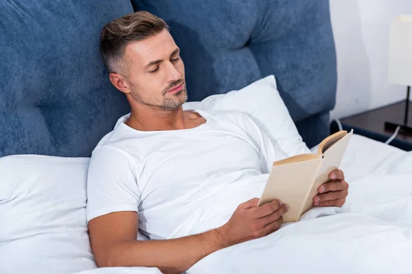 Adulto homem leitura livro enquanto deitado na cama em casa — Fotografia de Stock