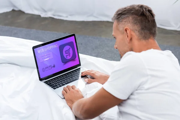 Rear view of adult man laying in bed and using laptop with web shopping on screen at home — Stock Photo