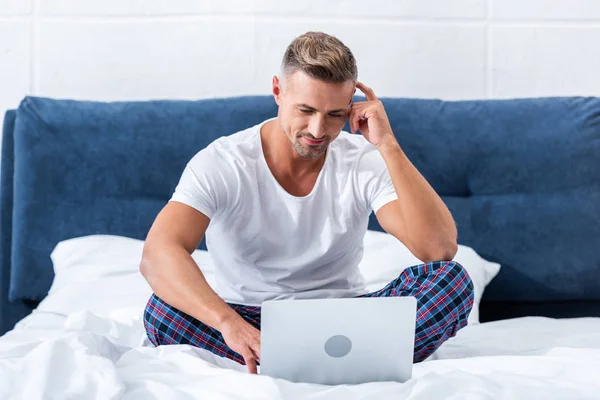 Handsome male freelancer sitting on bed and using laptop at home — Stock Photo