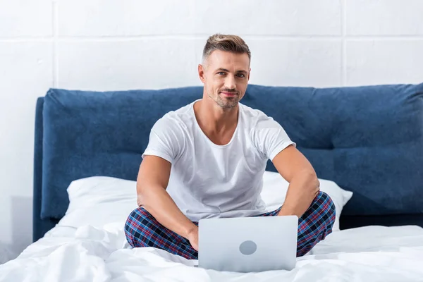 Sonriente freelancer masculino mirando a la cámara y sentado en la cama con el ordenador portátil en casa - foto de stock