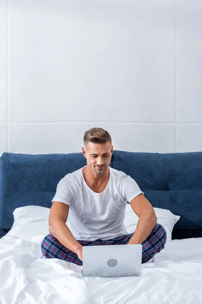 Adult male freelancer sitting on bed and using laptop at home — Stock Photo