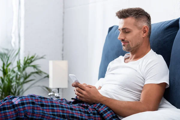Hombre alegre en camiseta blanca usando teléfono inteligente durante la mañana en la cama en casa - foto de stock