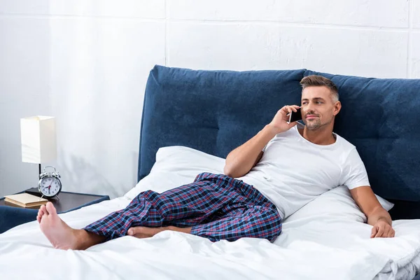 Cheerful adult man talking on smartphone while laying in bed at home — Stock Photo