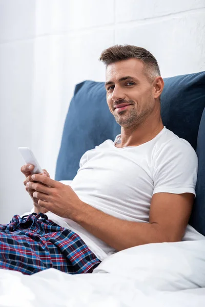 Усміхнений чоловік у білій футболці дивиться на камеру і використовує смартфон у ліжку вдома — стокове фото