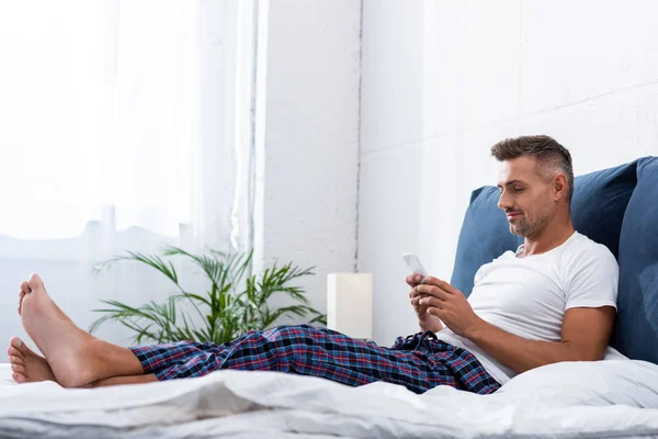 Hombre adulto en camiseta blanca usando teléfono inteligente durante la mañana en la cama en casa - foto de stock