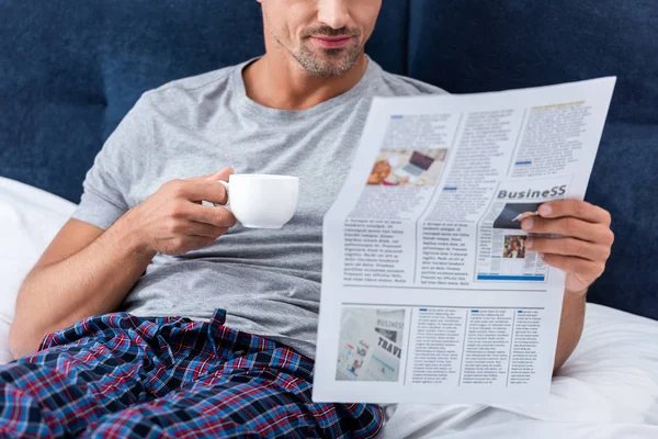 Imagen recortada de hombre de negocios con taza de café leer periódico de negocios en la cama en casa - foto de stock