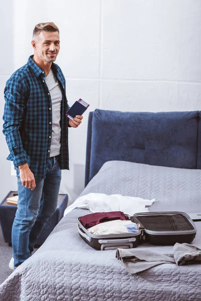 Happy adult man with passport and flight ticket standing near travel bag at home — Stock Photo