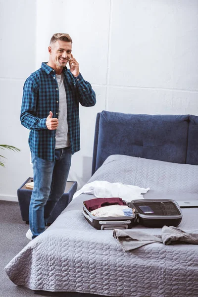 Sonriente hombre adulto hablando en el teléfono inteligente y haciendo gesto de pulgar hacia arriba cerca de equipaje en casa - foto de stock