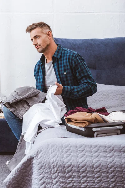 Вдумчивый мужчина сидит с одеждой рядом с колёсной сумкой в спальне дома — стоковое фото